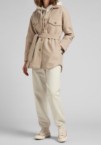 Persona vistiendo un abrigo beige con cinturón sobre una sudadera crema, pantalones jogger blancos y zapatillas altas beige, sobre un fondo gris liso.