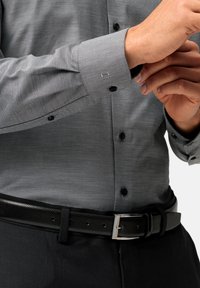 Grau gemusterte Hemd mit langen Ärmeln, schwarzen Knöpfen und geknöpftem Umschlag, kombiniert mit einem schwarzen strukturierten Gürtel und dunklen Hosen.