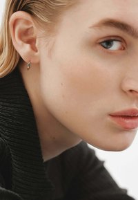 Pendiente en tono dorado con una pequeña gema de turquesa, mostrado en un primer plano de la oreja de un modelo. Diseño ligeramente curvado con un acabado brillante.