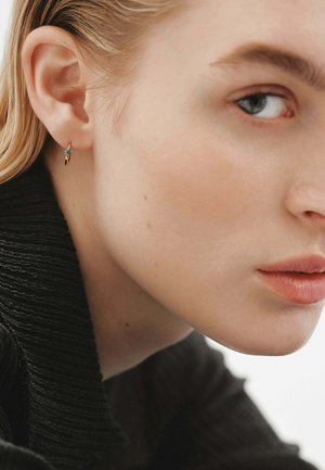Pendiente en tono dorado con una pequeña gema de turquesa, mostrado en un primer plano de la oreja de un modelo. Diseño ligeramente curvado con un acabado brillante.