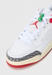 Biały sneaker z teksturowanej skóry, czerwonymi akcentami, zielonym plastikowym detalem, szarym wzorem na nakładce oraz czerwonym podeszwa. Posiada okrągłe sznurówki i logo.