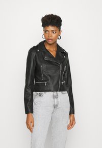 Schwarze Kunstleder-Cropped-Jacke mit Reißverschluss-Taschen und Schulterklappen; verfügt über einen breiten Kragen und kontrastierende silberne Hardware.