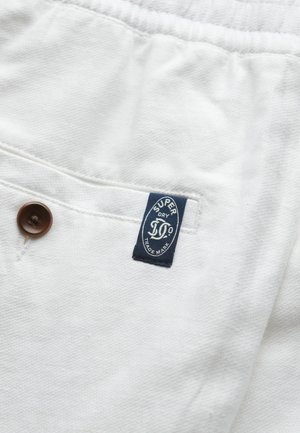 Fehér szövet, texturált mintával, egy barna gombbal és egy kék címkével, amelyen a "SUPERDRY" felirat és a védjegy adatai találhatók.