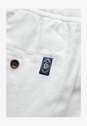 Fehér szövet, texturált mintával, egy barna gombbal és egy kék címkével, amelyen a "SUPERDRY" felirat és a védjegy adatai találhatók.