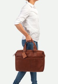 Bruine leren tote bag met dubbele handgrepen, een voorvak met rits en een bagagelabel. Gladde textuur met een minimalistisch ontwerp.