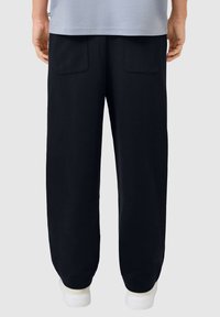 Persoon in zwarte losse broek met achterkant zakken en witte sneakers, staand tegen een effen lichtgrijze achtergrond.