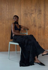 Zwarte jurk met textuur in bloemmotieven, gecombineerd met een zwarte top en elegante zwarte schoenen. Het model zit op een lichtgroene stoel tegen een houten achtergrond.