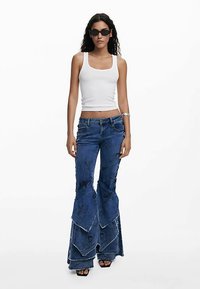 Jeans med høj talje og brede ben i mørkeblå denim, med lagdelt design og slidte detaljer. Kombineret med en tætsiddende hvid tanktop.