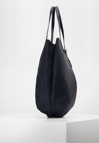 Mörkblå läderhandväska med en mjuk, texturerad yta; har två långa remmar och en rundad profil, vilande på en vit yta.