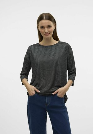 Maglia nera metallizzata a maniche lunghe con scollatura tonda e texture a coste, abbinata a jeans blu. Il modello sta in piedi con le mani in tasca.