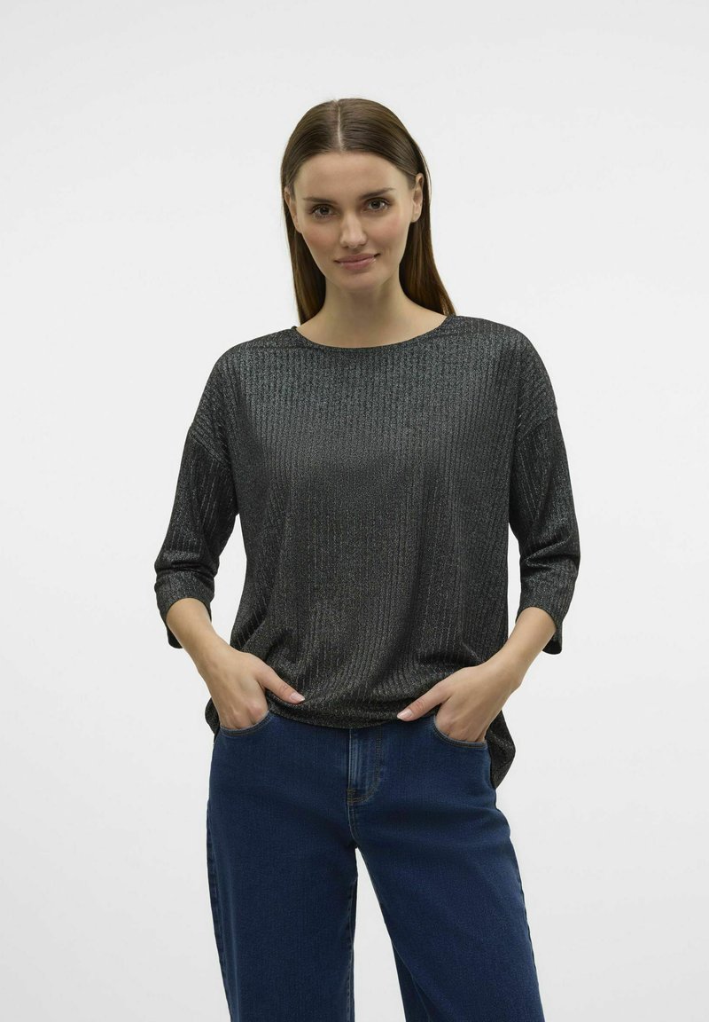 Maglia nera metallizzata a maniche lunghe con scollatura tonda e texture a coste, abbinata a jeans blu. Il modello sta in piedi con le mani in tasca.