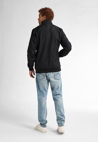 Giubbotto bomber nero con colletto stand-up, polsini a coste e finitura liscia, abbinato a jeans azzurri chiari e sneakers bianche.