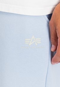 Világoskék pulóver, amely puha szövettextúrával rendelkezik, és egy fehér logója van, amely egy absztrakt „A” dizájnt ábrázol. Látható kéz pihen a ruhadarabon.