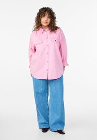 Rozā pogājama krekla ar divām krūšu kabatām, garām piedurknēm, kas satīti uz augšu, kopā ar platām zilām džinsa biksēm. Gluda auduma tekstūra.