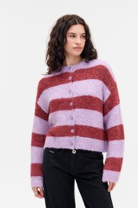 Donna che indossa un cardigan a righe lavanda e rosso ruggine con bottoni e pantaloni neri, in piedi contro uno sfondo chiaro e semplice, guardando a destra.