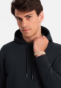 Felpa nera con cappuccio e lacci neri; modello che indossa un bracciale beige testurizzato al polso, mostrando un primo piano dei dettagli del tessuto e della vestibilità.