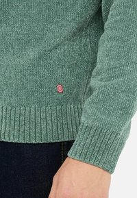 Maglione verde con texture, caratterizzato da polsini e orlo a coste, e un piccolo tag con logo rosa sul lato inferiore. Materiale morbido e tessuto.