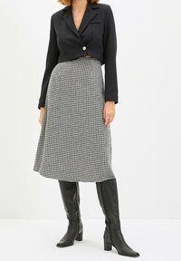 Czarna krótka marynarka z jednym guzikiem, połączona z czarno-białą spódnicą w desenie houndstooth sięgającą do kolan. Modelka nosi czarne kozaki za kolano.