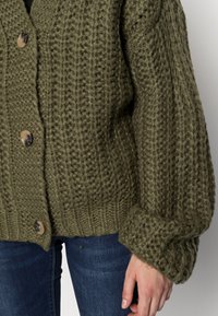 Objemný svetle olivově zelený pletený cardigan s velkými knoflíky, nošený přes modré džíny, s texturovanými ribovými a semennými úplety.