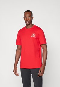 Camiseta de algodón roja con mangas cortas, cuello redondo y gráfico de "PREDATOR" en el pecho. Llevada con pantalones negros con rayas blancas.