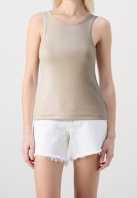Kvinna klädd i en beige ribbad ärmlös topp och vita fransade denimshorts, stående mot en neutral ljus bakgrund.