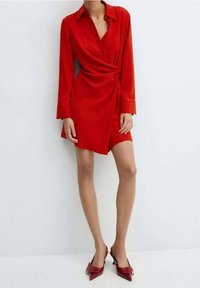 Vestido rojo envolvente con mangas largas, cuello tipo camisa y dobladillo asimétrico. Tela suave con un acabado elegante. Combinado con zapatos de tacón rojo puntiagudos.