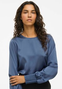 Vrouw met lang krullend haar draagt een blauwe satijnen blouse met geplooide mouwen, poseert met één arm gekruist over haar torso tegen een witte achtergrond.