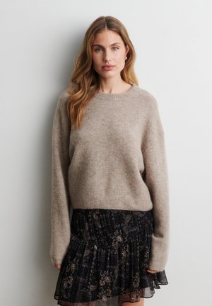 Femme aux longs cheveux châtains clairs portant un pull beige duveteux et une jupe noire à volants fleuris, debout contre un mur clair uni.
