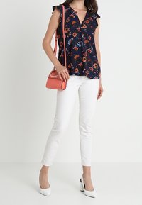 Marinblommig blus med volangärmar, ihop med vita byxor. Outfiten kompletteras av en korallfärgad handväska och vita högklackade skor.