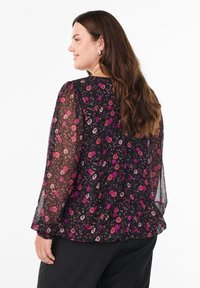 Frau mit langen braunen Haaren, die eine schwarze durchsichtige Bluse mit langen Ärmeln und einem roten und rosa Blumenmuster trägt, seitlich stehend und lächelnd.