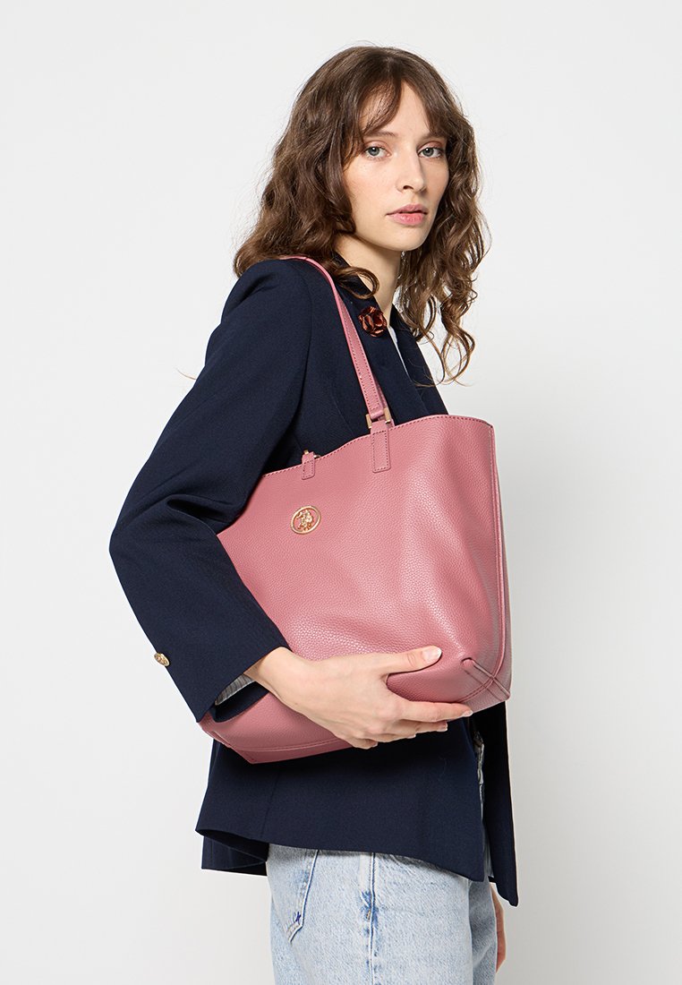 Sac en cuir rose avec une finition texturée, doté de deux poignées et d'un accent de logo rond, tenu par une personne portant un blazer bleu marine.