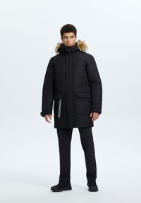 Parka nera con cappuccio foderato in pelliccia, dotata di chiusura a zip e bottoni a pressione, numerose tasche frontali e dettagli grigi sulla tasca sinistra.