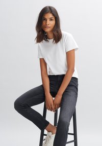 White crew-neck t-shirt paired with dark denim jeans. Model seated on a black stool, wearing white sneakers. Simple and clean design.