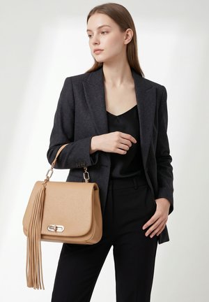 Giovane donna in abito nero che tiene una borsa in pelle color cuoio con dettagli a frangia lunga su sfondo bianco.