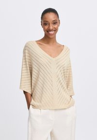 Beige pletený top s výstřihem do V, krátkými rukávy a diagonálním pruhovaným vzorem. Nosený s kalhotami světlé barvy, s jemným leskem.