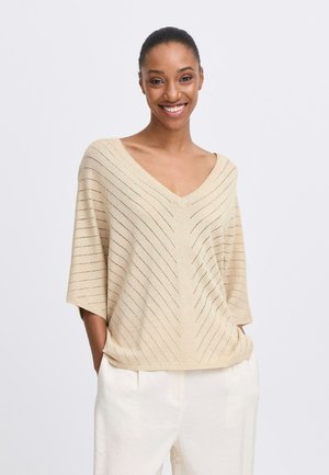 Beige gestrickter Oberteil mit V-Ausschnitt, kurzen Ärmeln und diagonaler Streifentextur. Getragen mit hellen Hosen, die einen subtilen Glanz aufweisen.