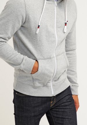Mann trägt hellgrauen Kapuzenpullover mit Reißverschluss und Kordeln sowie dunkle Jeans, die Hand in der Kapuzenjackentasche, steht vor einfarbigem Hintergrund.
