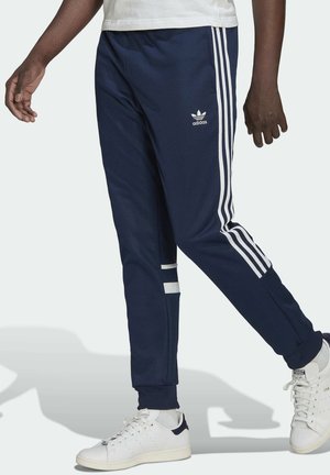 Persona che indossa pantaloni della tuta Adidas blu navy con strisce bianche e scarpe da ginnastica bianche, camminando con una gamba in avanti.