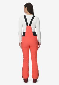 Koraalkleurige skibroek met zwarte verstelbare banden, slim-fit ontwerp en uitlopende pijpen. Voorzien van ritssluitingen bij de enkels en een rekbare tailleband voor comfort.