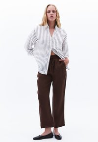 Camicia a righe con bottoni in bianco e nero, dalla vestibilità ampia, abbinata a pantaloni marroni con coulisse e ballerine nere. Design minimalista.
