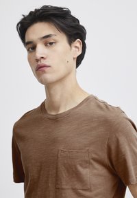 Hnědé tričko s krátkým rukávem vyrobené z měkké látky, s náprsní kapsou a kulatým výstřihem. Textura vypadá hladce a je lehká.