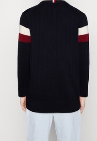 Granatowy sweter z prążkowanego dzianiny z białymi i czerwonymi paskami na górnych partiach rękawów, noszony z jasnoniebieskimi dżinsami, widziany od tyłu.