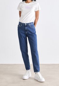 Bílá tričko s světle fialovou grafikou, spojené s modrými džíny s vysokým pasem, rovnými nohavicemi a předními kapsami. Bílé tenisky.