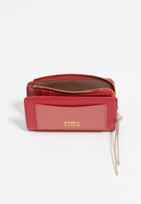 Cartera de cuero rojo con superficie texturizada, herrajes dorados y un panel rosa en contraste que presenta el nombre de la marca. Cierre con cremallera y correa para la muñeca.