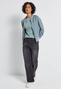 Helleblaue Bomberjacke, gestreiftes petrolfarbenes Oberteil, schwarze weite Jeans und weiße Sneaker. Die Jacke verfügt über Reißverschlusstaschen und einen gerippten Kragen.