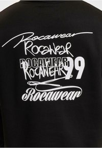 Zwarte katoenen sweatshirt met meerdere witte Rocawear-logo's in verschillende lettertypen en maten, inclusief het nummer 99, op de achterkant.