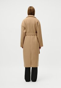 Cappotto beige in lana con vita cintrata, colletto oversize e maniche lunghe. Il cappotto ha una linea dritta e arriva sotto il ginocchio.