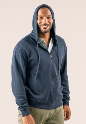 Dunkelblaues Zipper-Hoodie aus weichem Material, mit einer Kapuze mit Kordelzug, gerippten Bündchen und Fronttaschen. Darunter wird ein hellgrünes Shirt getragen.