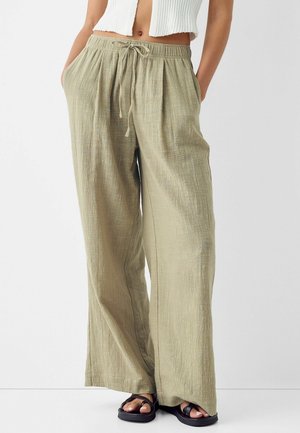 Mujer con pantalones de lino beige de corte holgado con cintura ajustable, top blanco corto de punto y sandalias negras con tiras cruzadas.