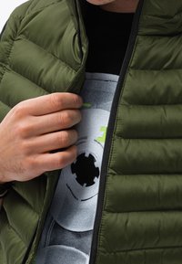 Oliwkowa pikowana kamizelka z zamkiem błyskawicznym z przodu, z poziomymi przeszyciami i czarnym zamkiem. Pod spodem szara koszulka z grafiką z okrągłymi wzorami.